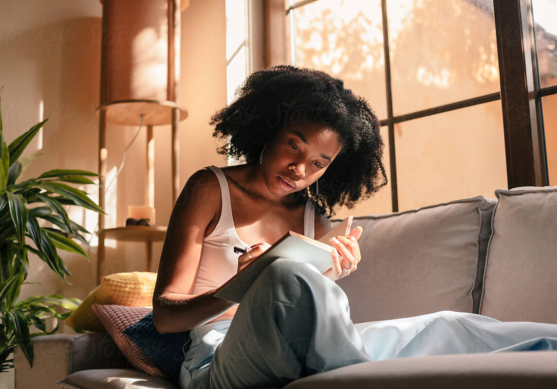 Young black woman at home, sitting on the couch and writing in her notebook.